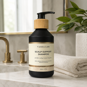 A bottle of Vassia Labs Scalp Support Shampoo stands on a marble countertop in a bathroom setting, with a folded towel, brass faucet, and green plant in the background.